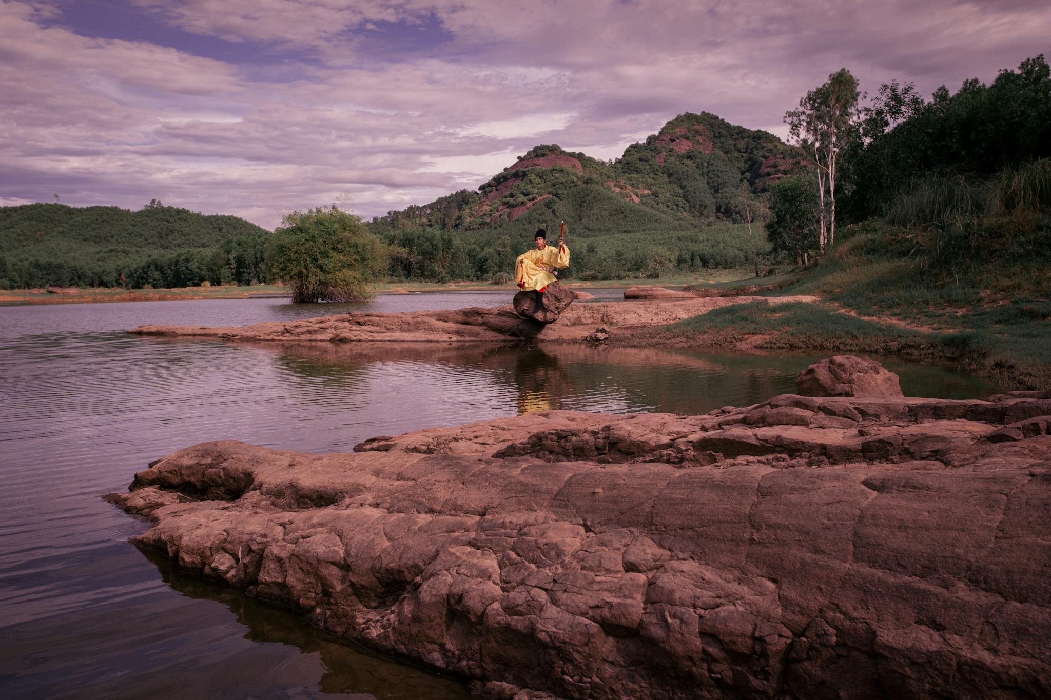 A solitary figure meditates by a tranquil river in Hội An, Vietnam's lush countryside.